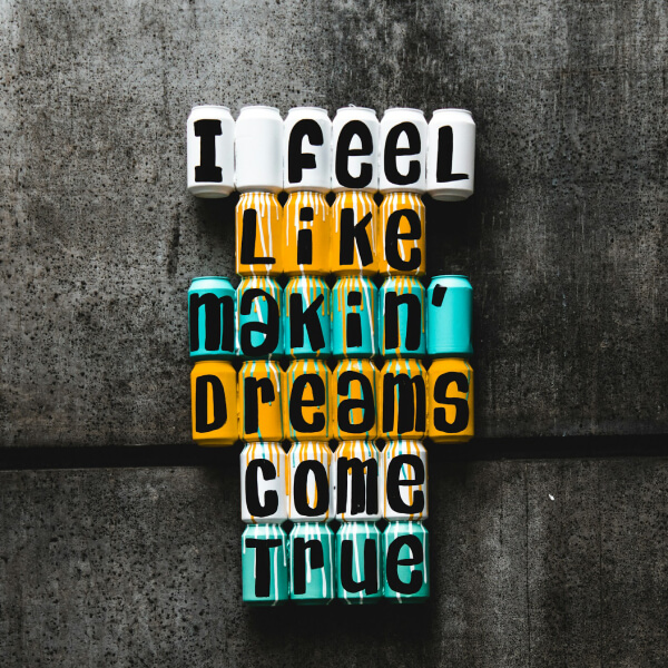 The phrase 'I feel like makin' dreams come true' is written in bold marker pen on painted drink cans. The pen is coloured black and the cans alternate in rows coloured white, orange and turquoise. They lie on an industrial concrete background.