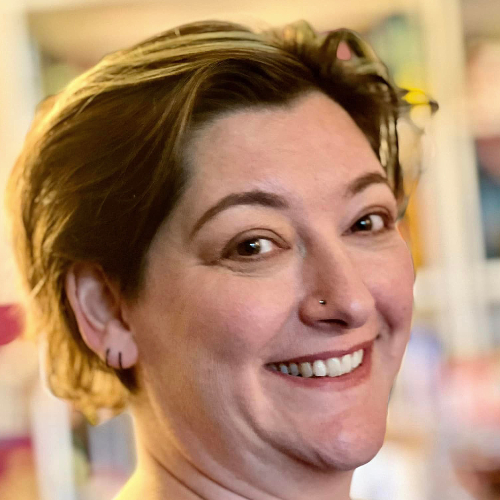 Photo of Emma Leadley, a white non-binary person taken side on. The background is blurred and their face is in sharp focus. They are smiling at the camera and they have crow's feet at the corners of their eye. Their hair is brown with a paler streak and they have multiple ear piercings and a nose piercing visible.
